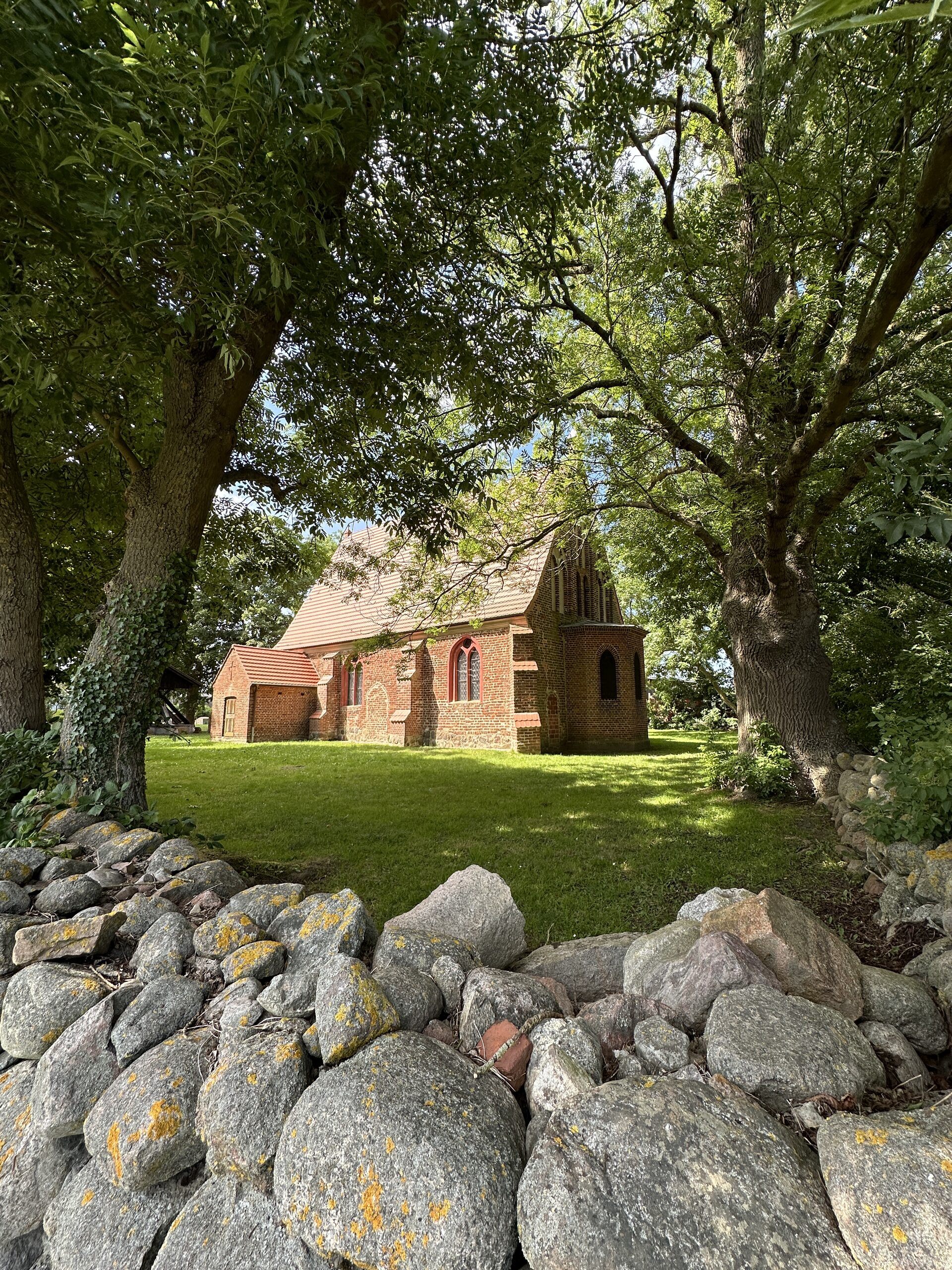 Kleine Backsteinkirche zwischen alten Bäumen hinter einer Feldsteinmauer