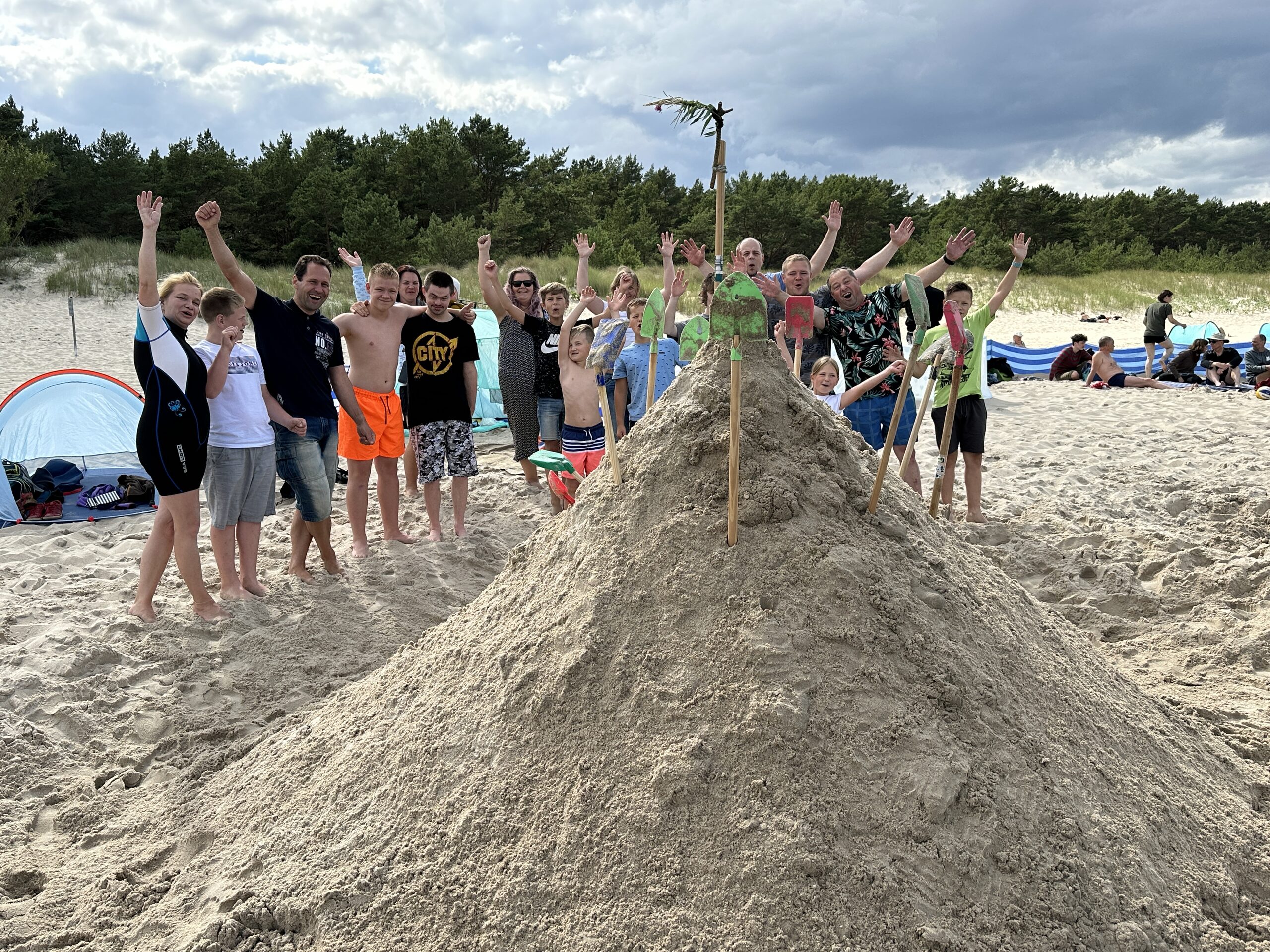 Gruppe mit erhobenen Armen hinter einer großen Sandburg am Strand