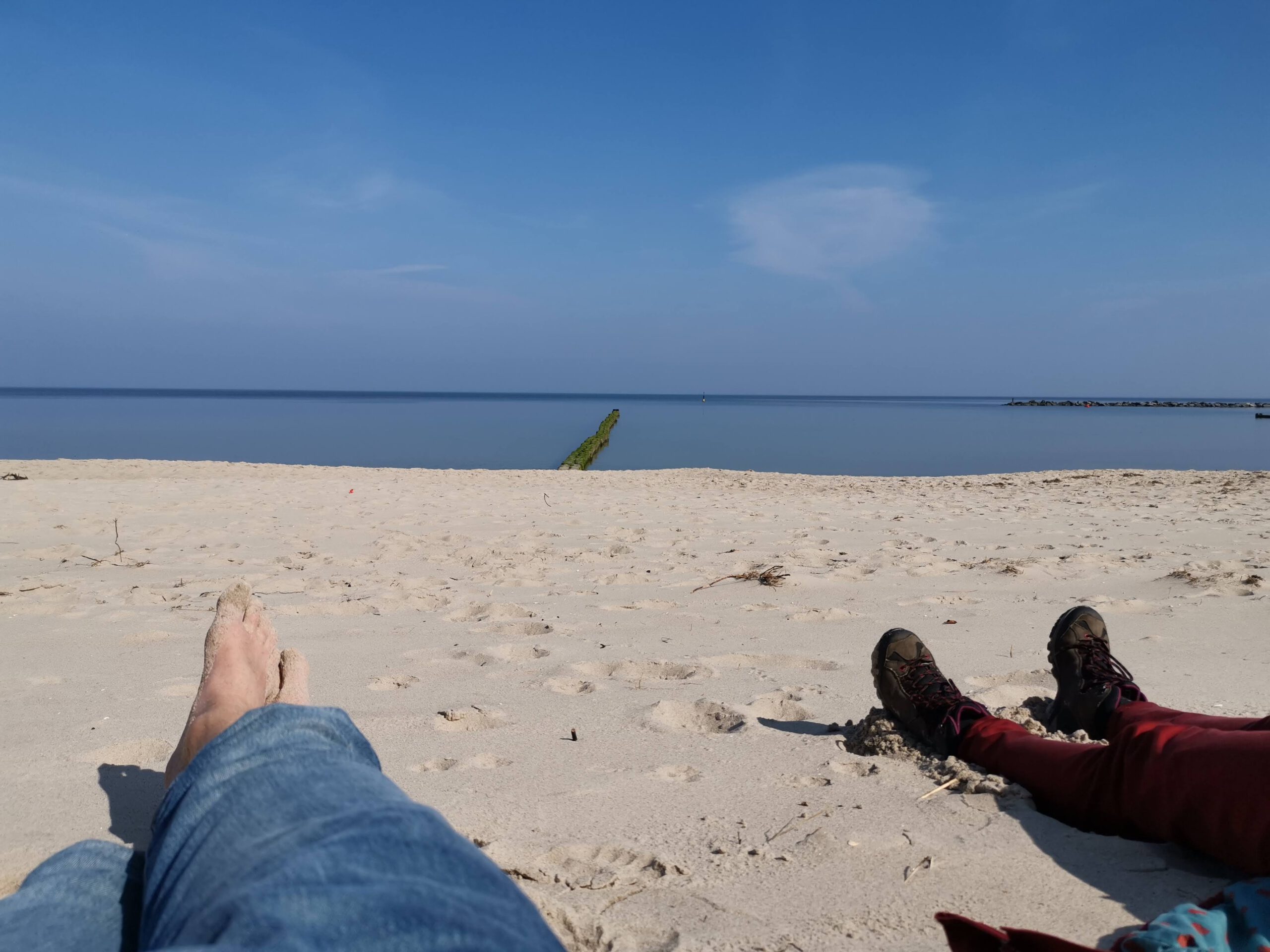 Füße im Sand am Ostseestrand bei blauem Himmel, im Hintergrund eine Buhne im Wasser