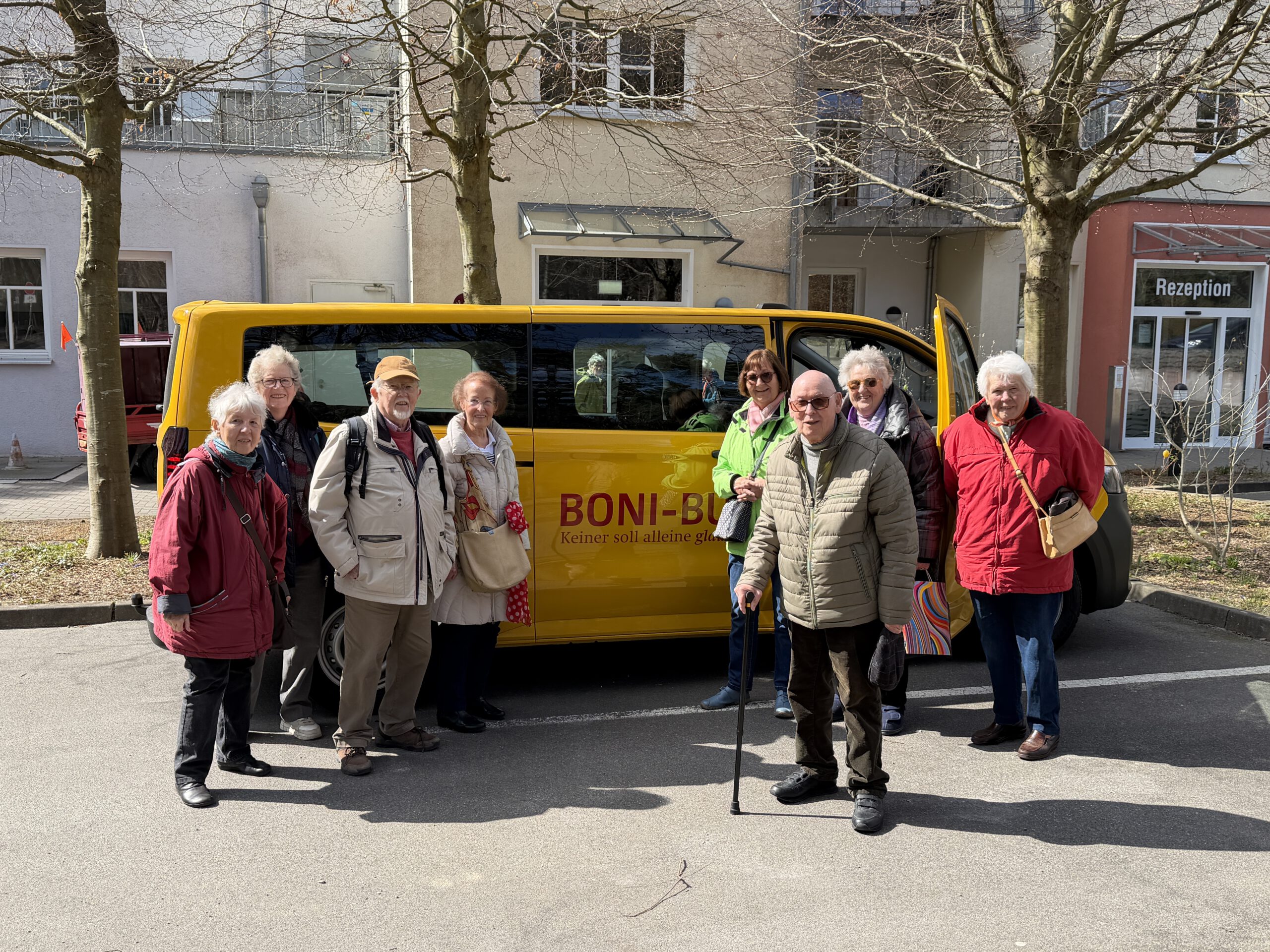 Gruppe von Senioren posiert lächelnd vor einem gelben Boni-Bus Kleinbus vor der Rezeption